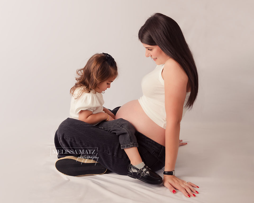 mom sitting against a white backdrop with toddler in her lap both looking at each other during their maternity session in metro detroit