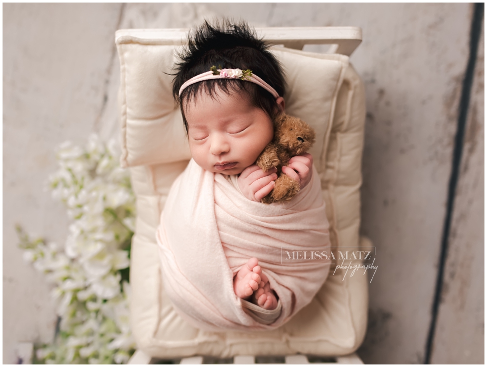 Newborn baby girl wrapped in pink swaddle surrounded by soft white floral arrangement in Shelby Township studio.