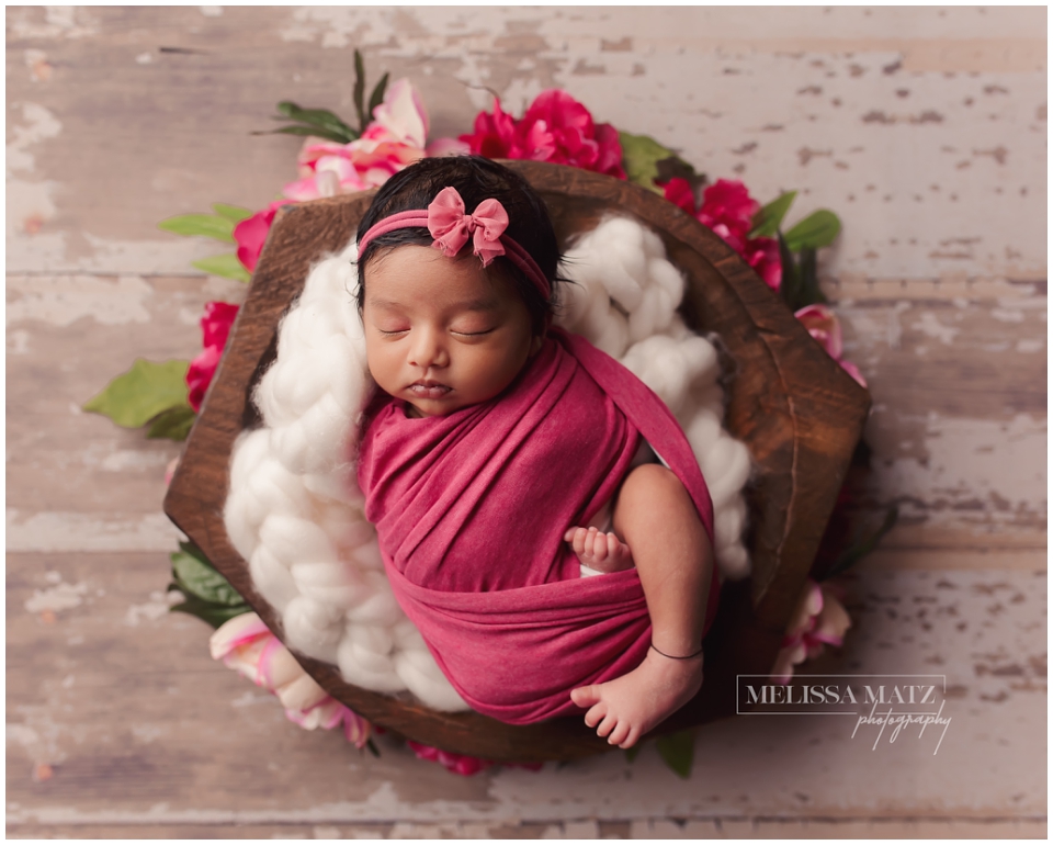 Newborn baby girl wrapped in a cranberry swaddle surrounded by cranberry pink floral arrangement in Shelby Township studio