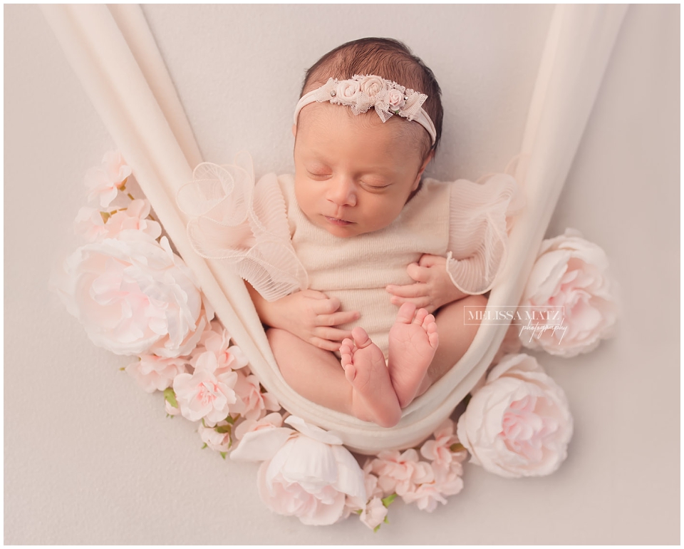 Sleeping newborn in an outfit resting on cream backdrop surrounded by white and pink flowers captured by Shelby Township MI newborn photographer.