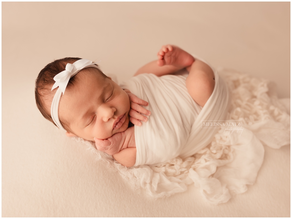 newborn baby swaddled in white wrap her hand cupping her cheek and her tiny foot peeking out