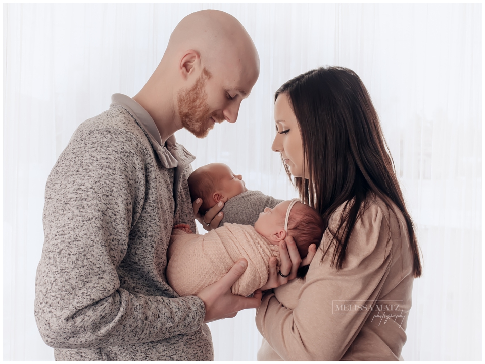parents holding their twin babies swaddle wrapped in front of window during their newborn photo session