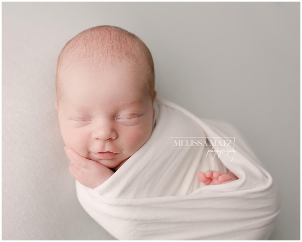 newborn baby swaddle wrapped in white smirking during his newborn session in shelby township mi photo studio