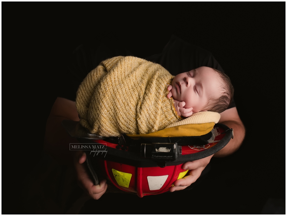 Newborn baby swaddled and sleeping in firefighter helmet held by dad dressed in black on a black backdrop during their newborn session in Shelby Township studio.