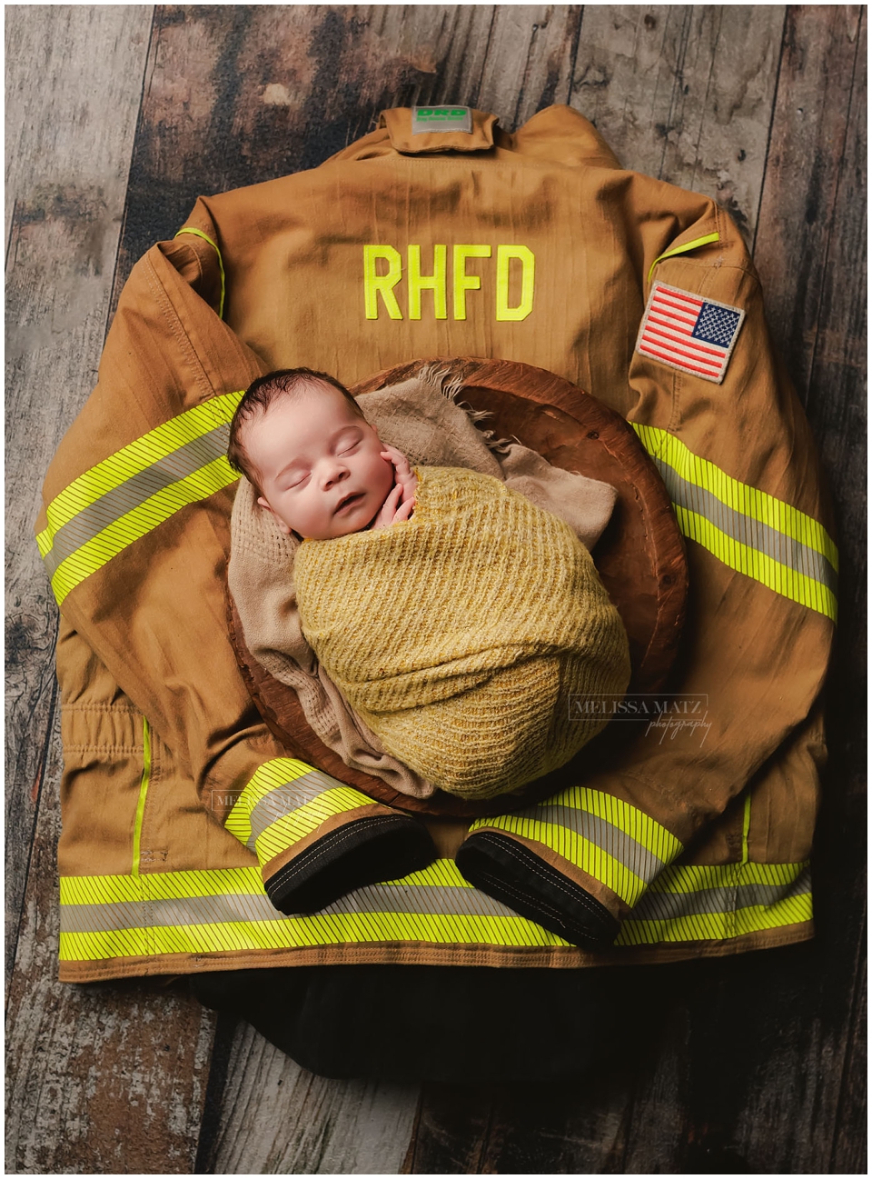 Swaddled newborn asleep in wooden bowl placed on father’s firefighter coat during his newborn session with Melissa Matz Photography
