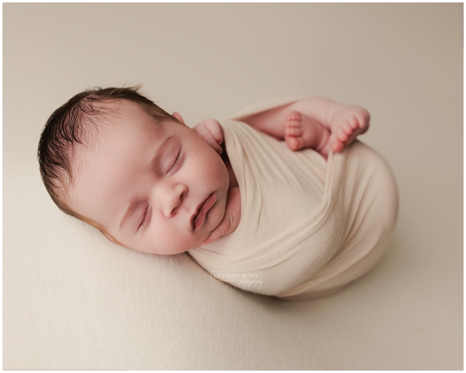 newborn baby swaddled in soft cream and posed with tiny hand cupping his chin
