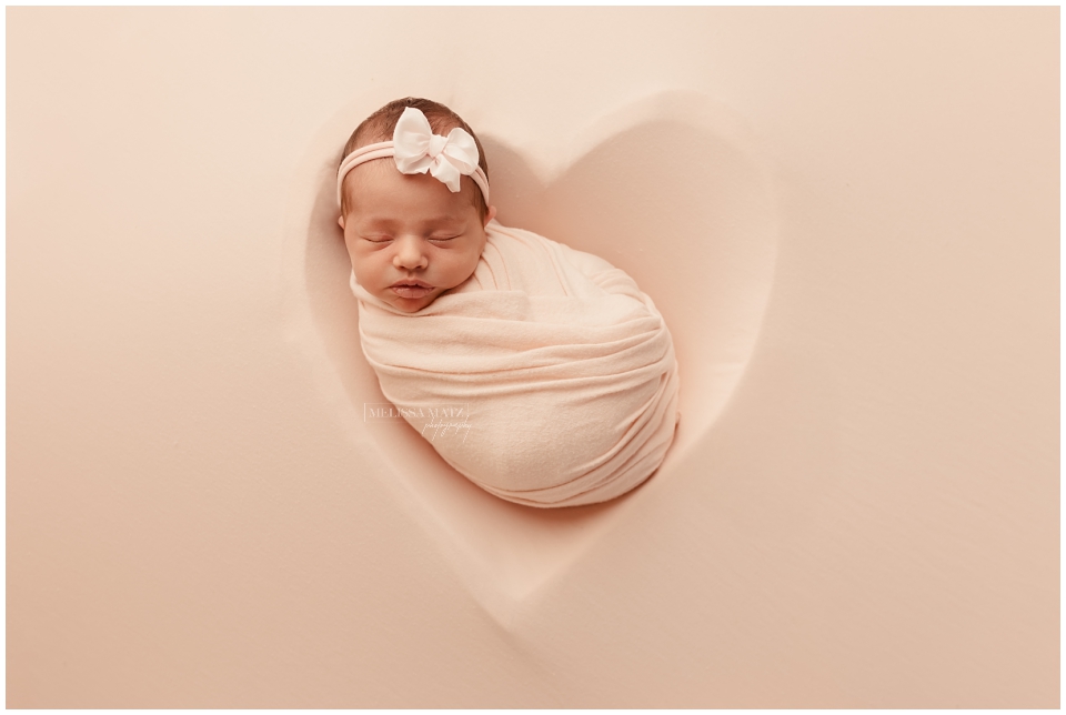 newborn baby with pink bow headband wrapped sleeping on a heart fabric