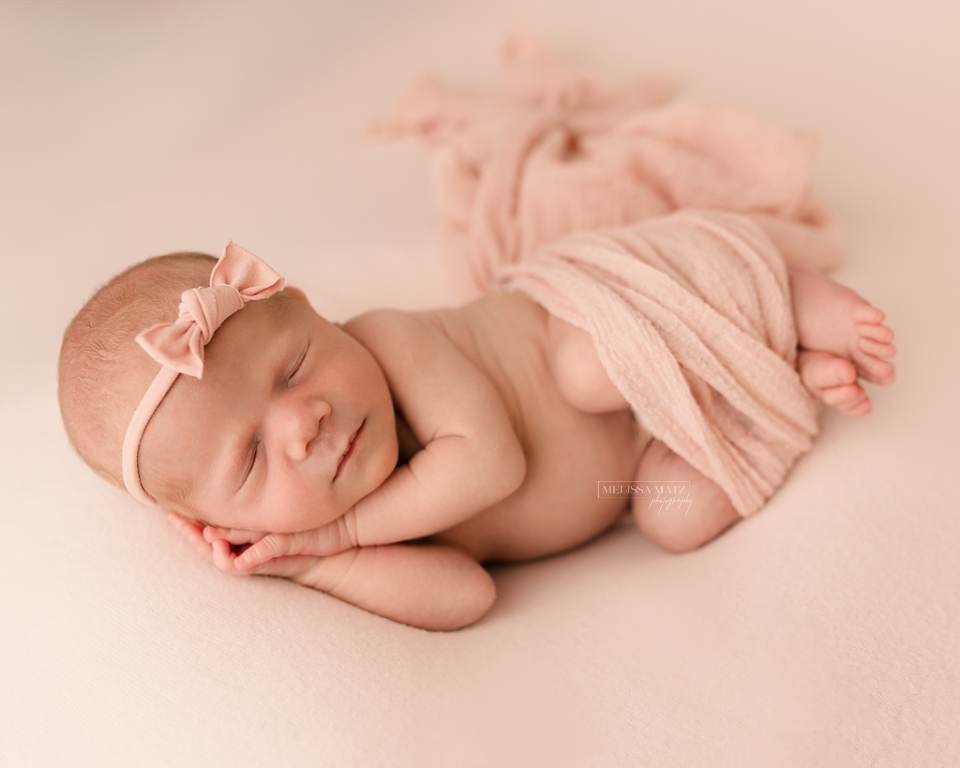 newborn baby sleeping hands under her cheek on blush pink fabric with tiny pink bow