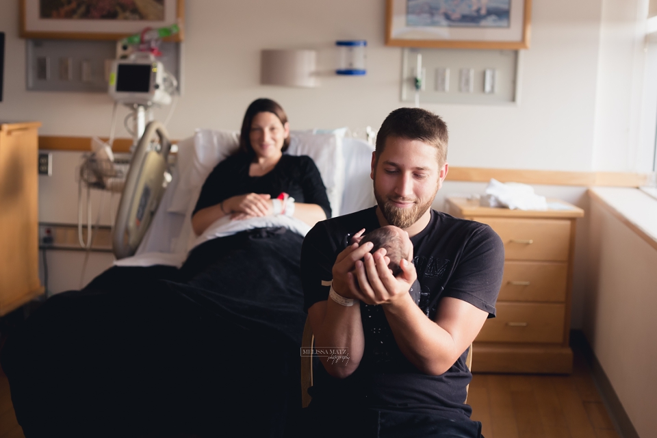 new father holding his brand new baby while mom looks on in there hospital bed