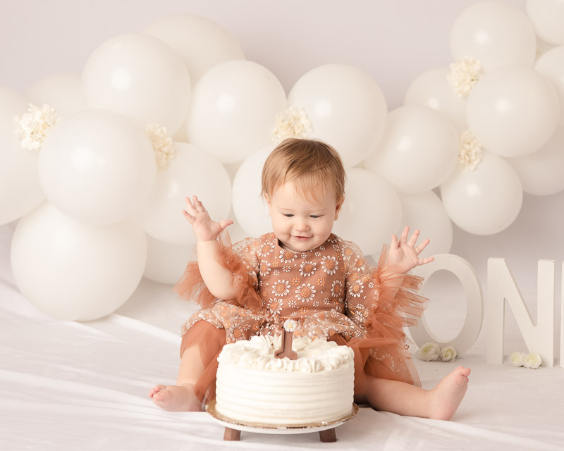 One year old baby girl in rust lace dress sitting in front of a white cake during a cake smash session at Melissa Matz Photography in Shelby Township