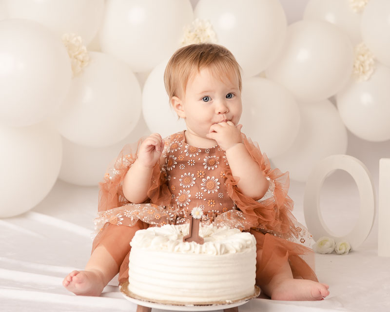 One year old baby girl in rust lace dress tasting cake in front of white balloon backdrop during a cake smash session at Melissa Matz Photography