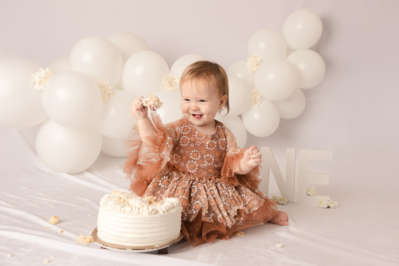 One year old baby girl laughing holding smashed cake pieces in front of white balloon backdrop during a cake smash studio session