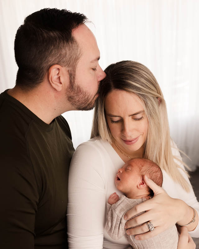 Both parents close together, dad kissing mom's temple, holding their newborn in a tender unscripted moment