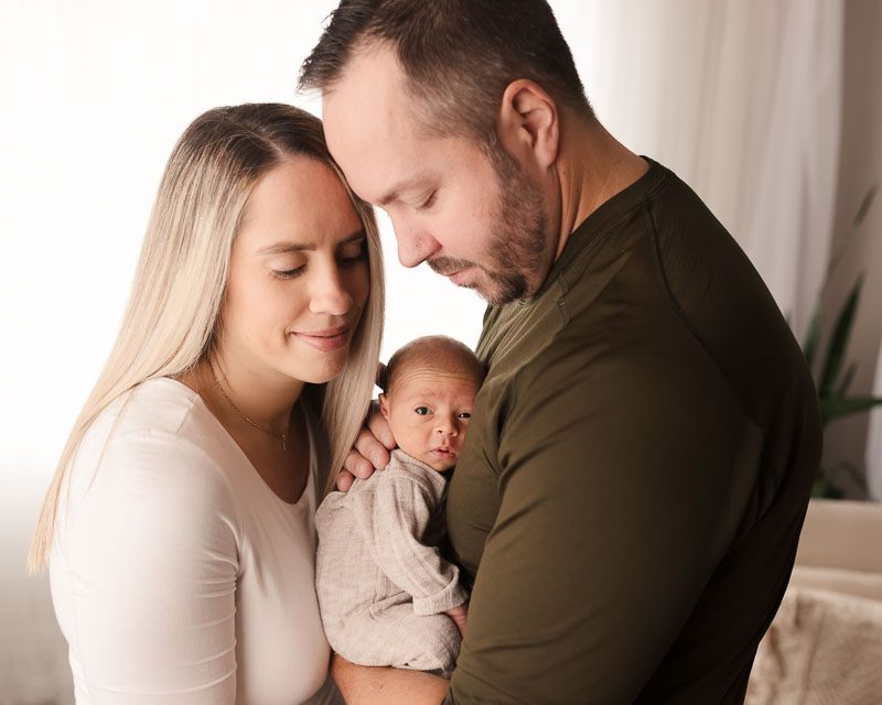 Mother and father standing together, both looking down at their newborn in a softly lit studio