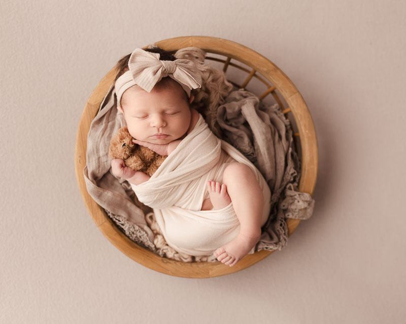 Newborn baby sleeping in studio basket with cream blanket, headband, and cradling a tiny teddy bear