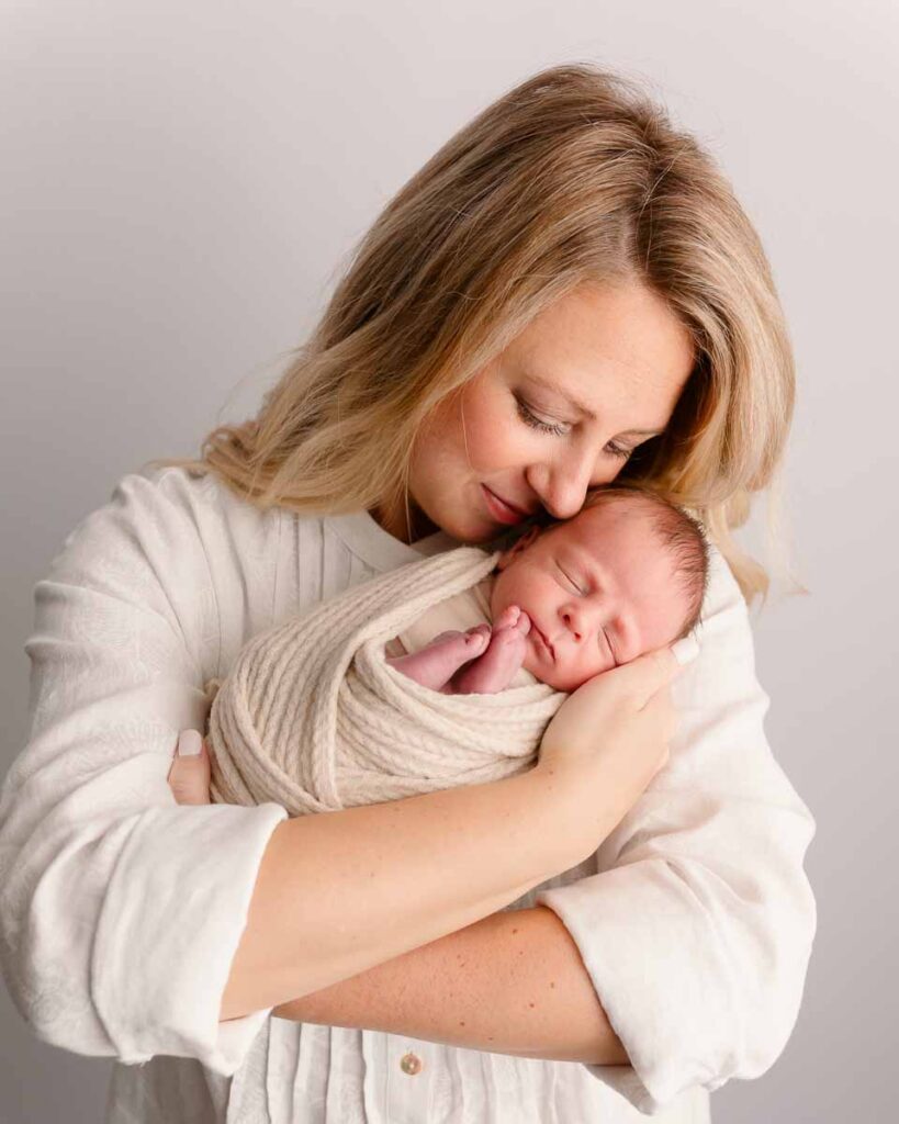 Mother in white holding her swaddled newborn baby with eyes closed during a studio newborn session at Melissa Matz Photography in Shelby Township Michigan