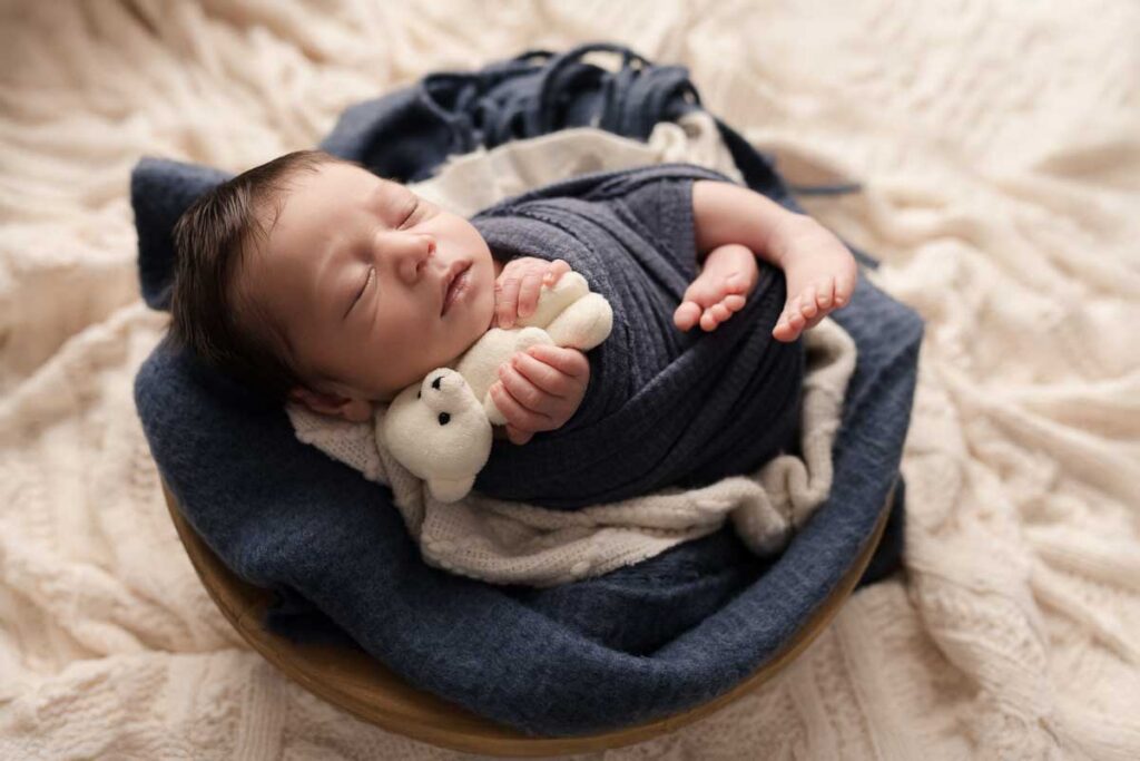 Newborn baby swaddled in a navy wrap with a stuffed bear during a professional newborn session in Shelby Township Michigan